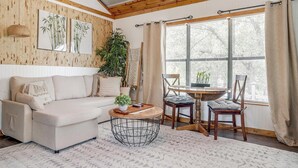 Ein gemütliches Wohnzimmer mit einer Couch, einem runden Couchtisch und einem Essbereich mit Stühlen. Ein großes Fenster mit Vorhängen, ein Topfpflanze und Kunstwerke an der Wand.