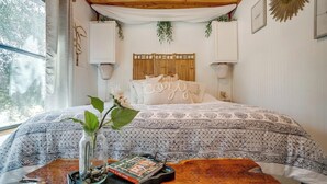 Ein Schlafzimmer mit einem Bett, einem Nachttisch mit Buch und Vase, einem Fenster mit Vorhängen und einer Holzbalkendecke.
