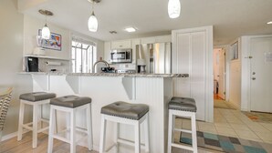 A kitchen with white cabinets, a microwave, a refrigerator, and two bar stools with cushioned seats. - Coastal Sun & Poolside Fun: Myrtle Beach Condo! (Myrtle Beach)