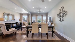 A spacious living room with a dining area, large windows, and a ceiling fan. - Beloved Haus - Hot-Tub, 4-Min to Main Street! (Fredericksburg)