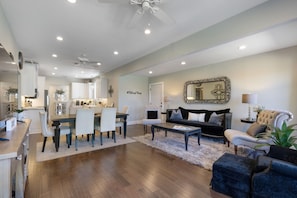A spacious living room with a dining area, a black sofa, a white armchair, and a large mirror.