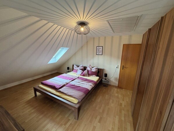 A bedroom with a sloped ceiling, a bed with a wooden frame, a bedside table, a wall-mounted light fixture, and a door.
