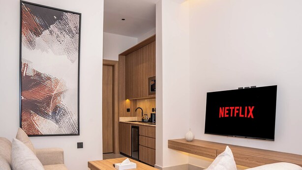 Un salon moderne doté d’un téléviseur à écran plat affichant le logo « NETFLIX », d’un coin repas avec une table en bois et d’une cuisine avec des meubles en bois.