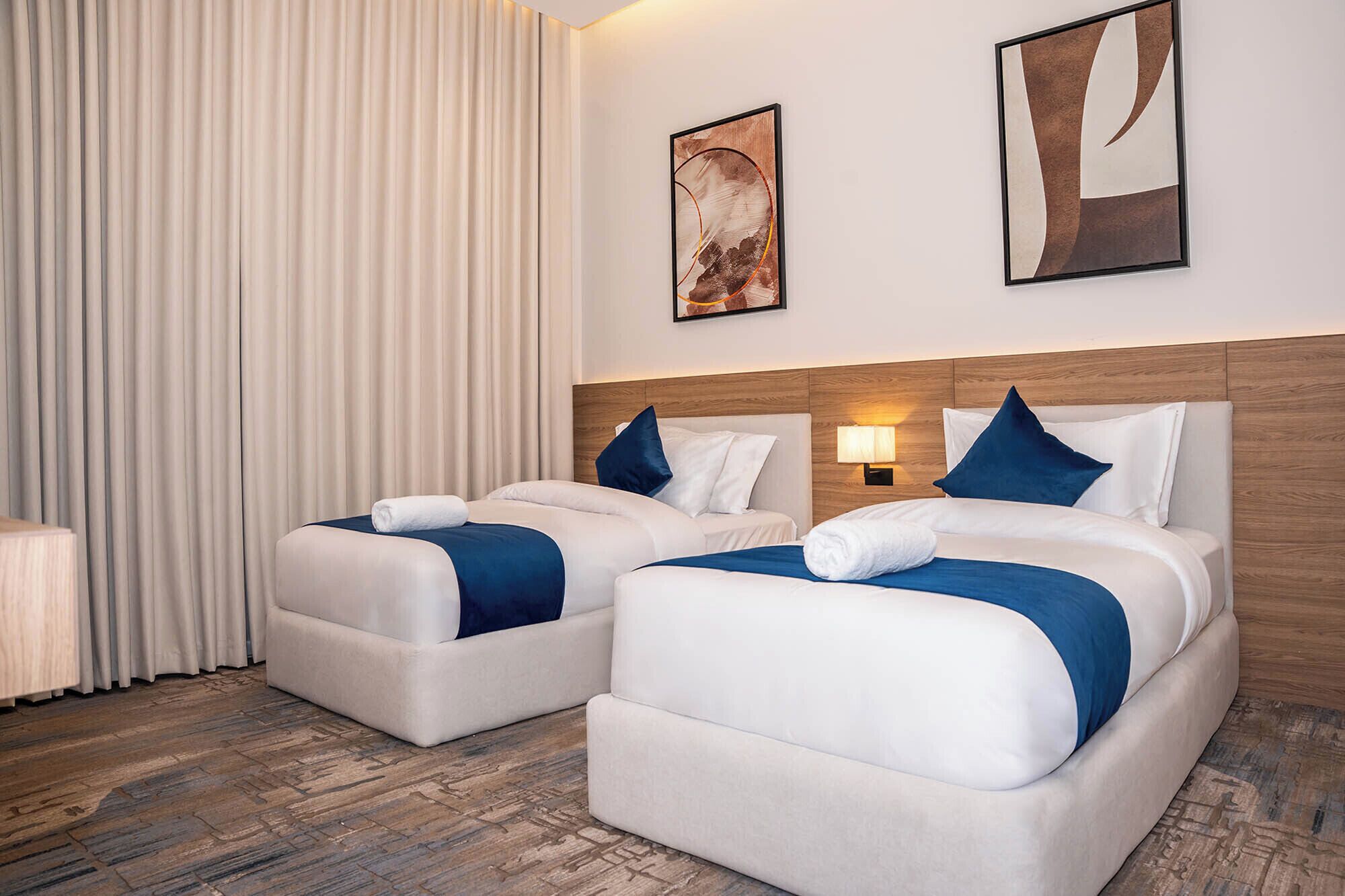 Une chambre d’hôtel avec deux lits, une tête de lit en bois, une table de chevet, une lampe et une grande fenêtre avec des rideaux.