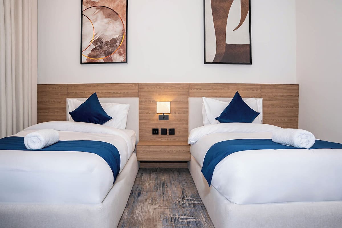 Hotellihuone, jossa on kaksi sänkyä, puinen pääty ja seinään kiinnitetty valaisin sekä taideteoksia.