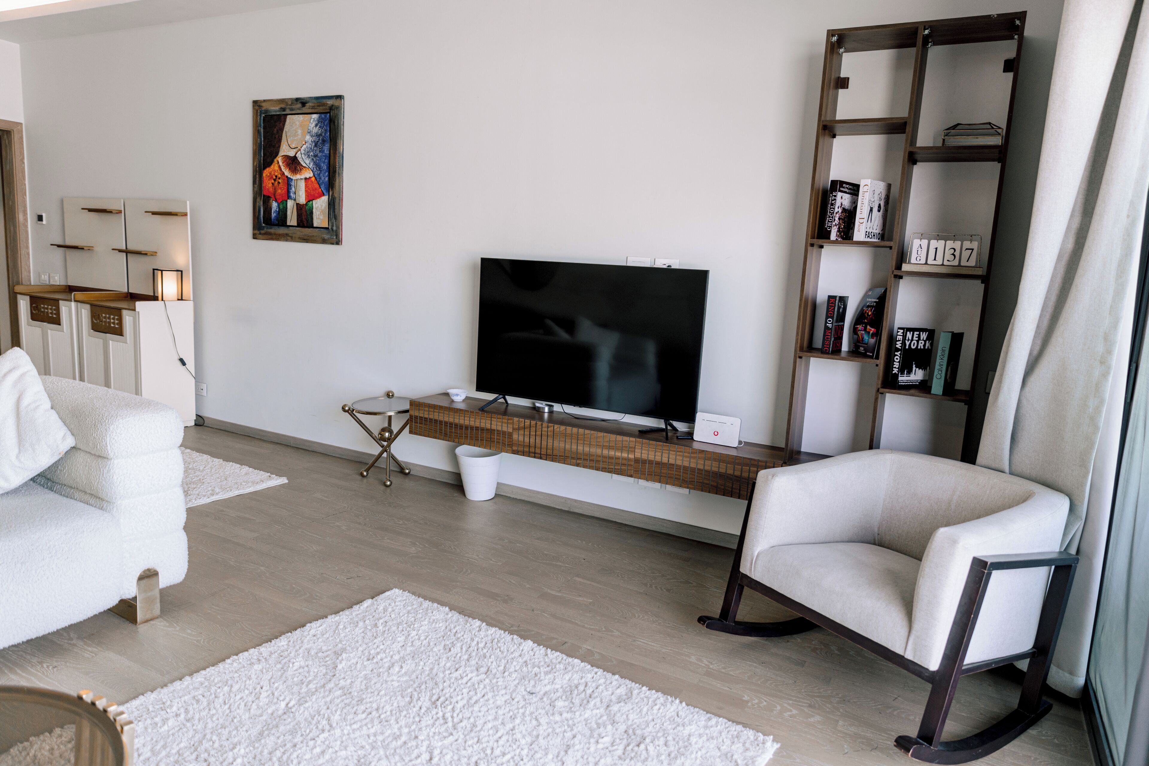 Uma sala de estar moderna com uma televisão de tela plana, uma estante, um sofá branco e uma mesa de centro.