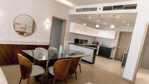 A modern kitchen with white cabinets, a central island, and a dining area with a glass table and wooden chairs. - Alsawah - Skyline 3BR - Aeon Towers (6th of October City)