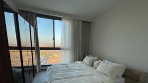 A bedroom with a large bed, a view of a cityscape through a window, and a balcony. - Alsawah - Skyline 3BR - Aeon Towers (6th of October City)