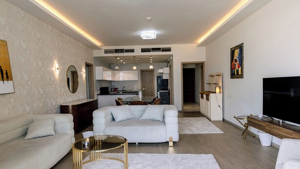 A modern living room with a sofa, coffee table, and TV. There is a kitchen in the background. - Alsawah - Skyline 3BR - Aeon Towers (6th of October City)