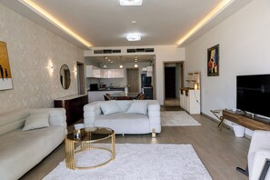 A modern living room with a sofa, coffee table, and TV. There is a kitchen in the background. - Alsawah - Skyline 3BR - Aeon Towers (6th of October City)