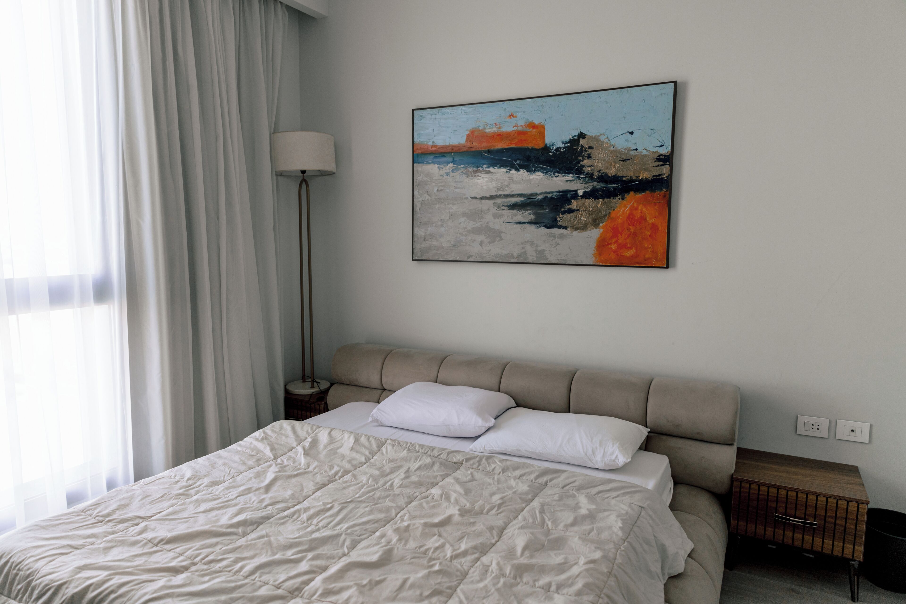 Ein ordentlich bezogenes Bett mit einem beige gefärbten Kopfteil, einem weißen Kissen und einer weißen Daunendecke. Über dem Bett hängt ein Landschaftsbild.