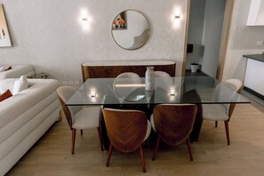 A modern dining area with a glass table surrounded by wooden chairs, a round mirror on the wall, and a white sofa in the background. - Alsawah - Skyline 3BR - Aeon Towers (6th of October City)