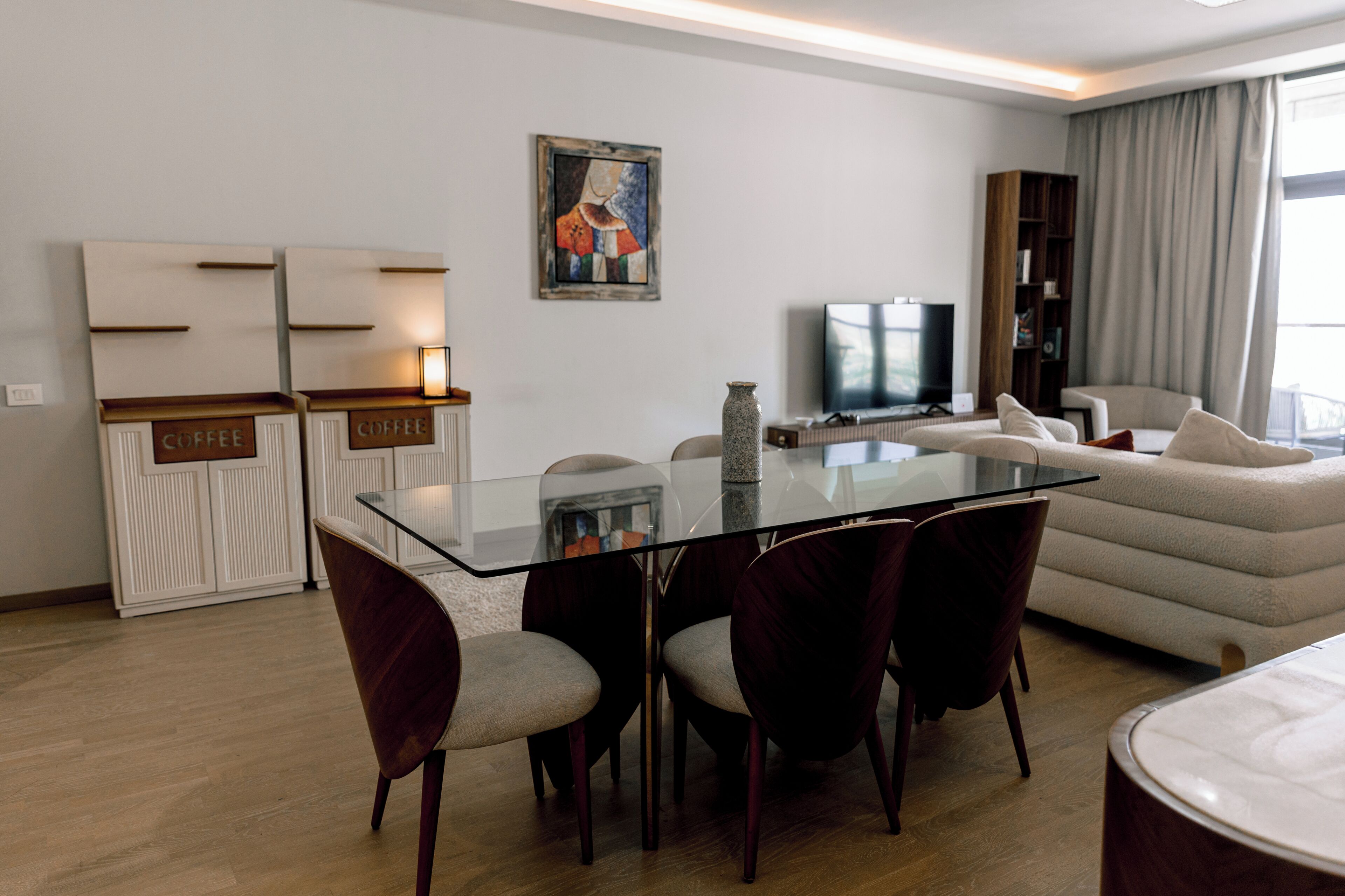 A modern living room with a glass dining table, dark chairs, a sofa, a television, and a bookshelf.