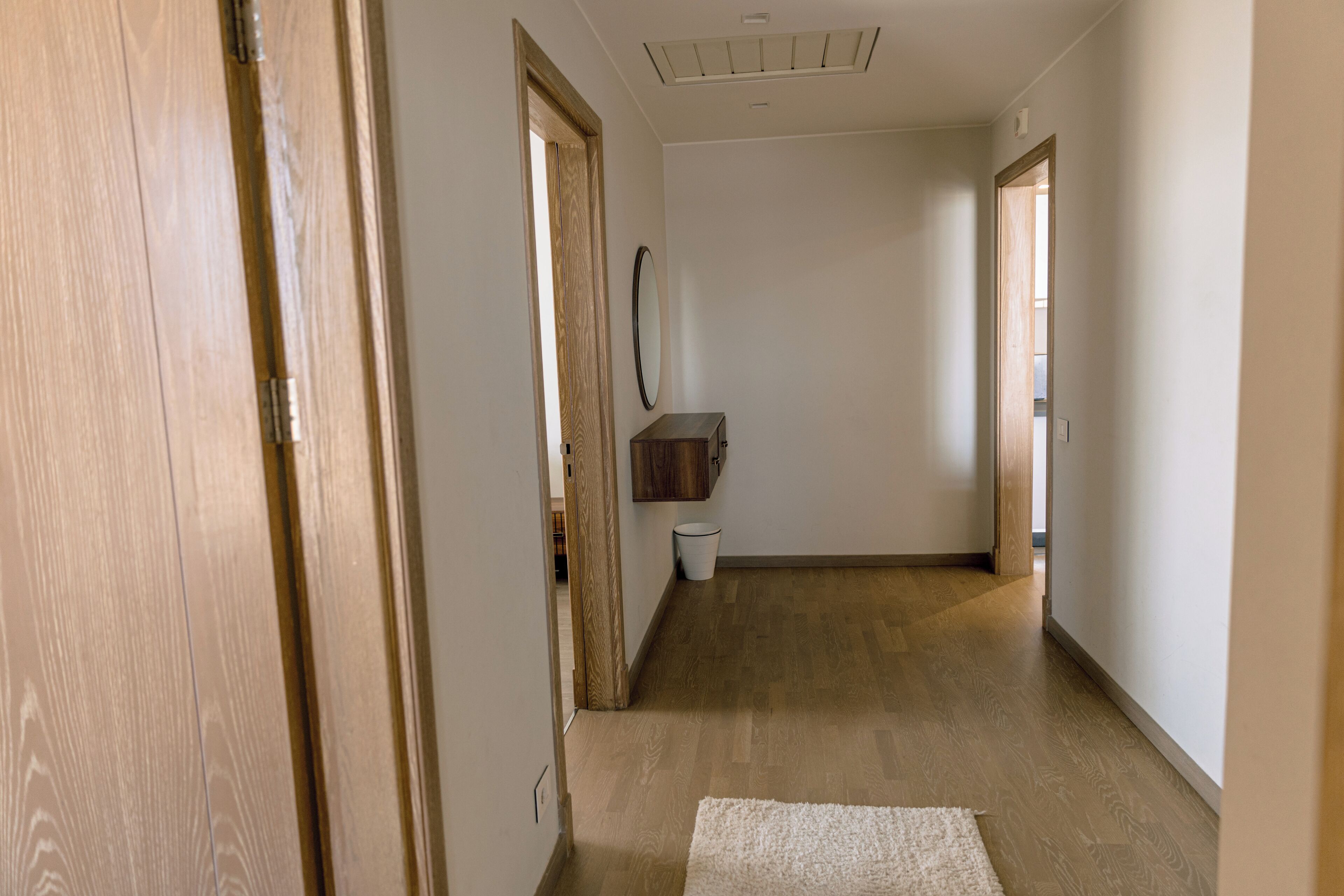 A hallway with wooden flooring, a white rug, and a wooden door on the left.