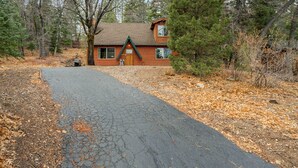 A log cabin with a paved driveway, surrounded by trees and a dry leaf-covered ground. - Felthaus Family Cabin (Big Bear Lake)