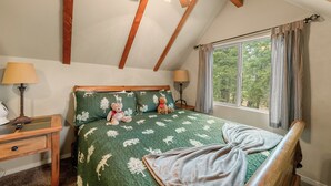 A bedroom with a sloped ceiling, a bed with a green quilt, two bedside lamps, a window with curtains, and a wooden nightstand with a lamp. - Felthaus Family Cabin (Big Bear Lake)