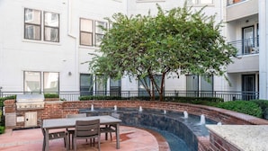 Une terrasse extérieure avec un mur en briques, un barbecue, une table avec des chaises et un élément décoratif aquatique.