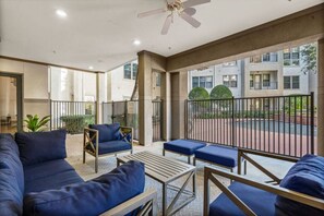 Een overdekt terras met banken en tafels, een plafondventilator, en uitzicht op een gebouw en straat.