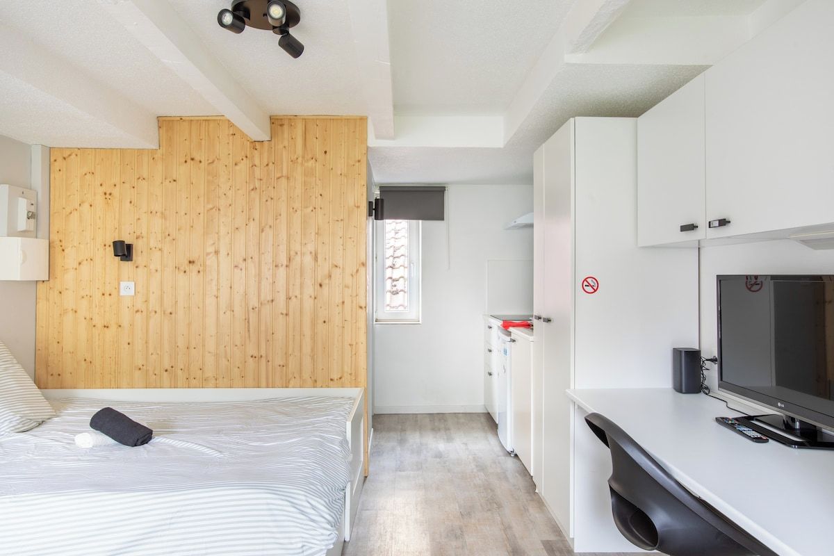 Une chambre à coucher avec une tête de lit en bois, un lit recouvert d’une couvre-lit à rayures, une table de chevet avec une lampe et un téléviseur posé sur une commode blanche.