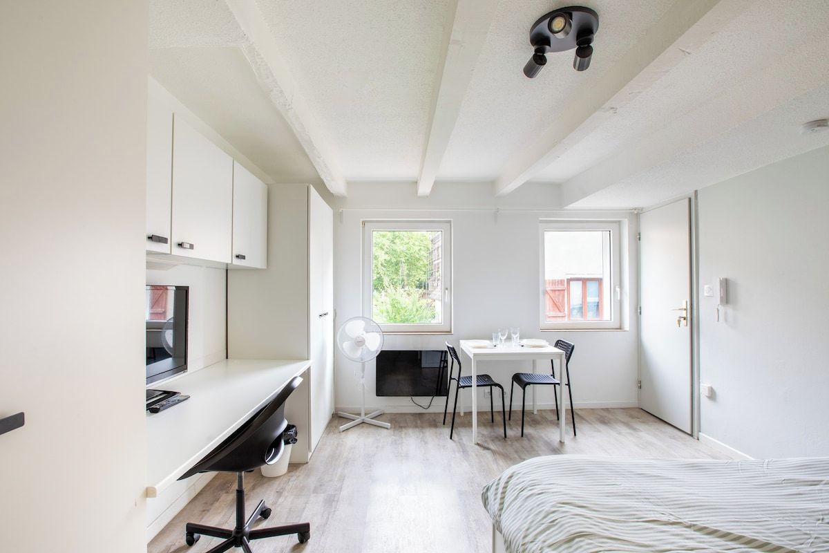 Een kleine kamer met een bed, een bureau, een stoel, een tafeltje en een raam.