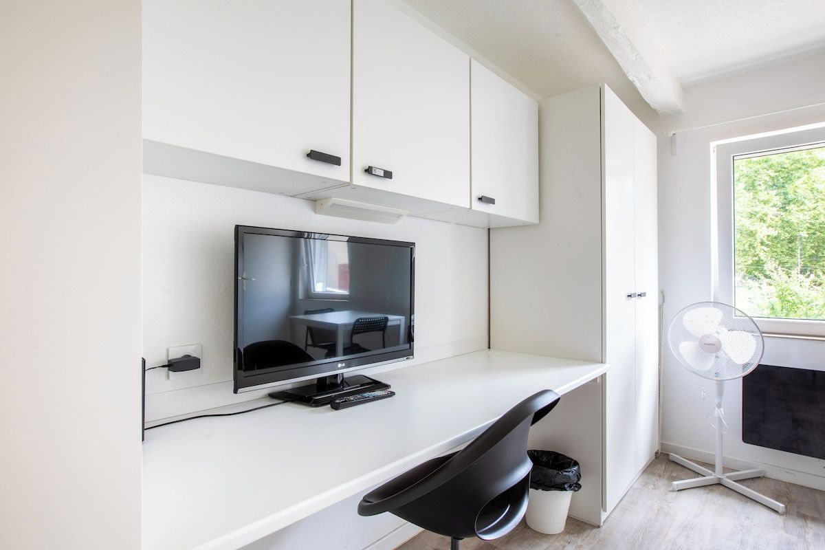 Habitación moderna con una televisión de pantalla plana sobre un escritorio blanco, una silla negra, un ventilador blanco y una ventana con vistas a zonas verdes.