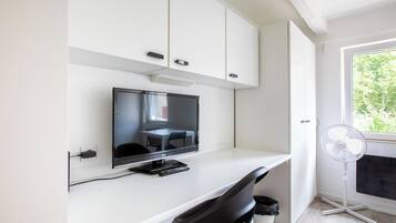 Habitación moderna con una televisión de pantalla plana sobre un escritorio blanco, una silla negra, un ventilador blanco y una ventana con vistas a zonas verdes.