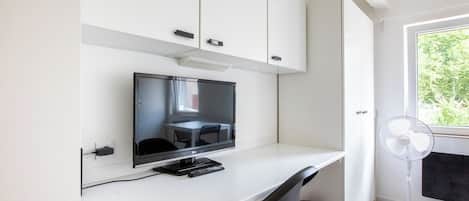 A modern room with a flat-screen TV on a white desk, a black chair, a white fan, and a window with a view of greenery.