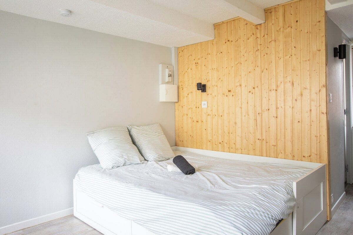 Una cama individual con ropa de cama a rayas, un panel de pared de madera, una mesita de noche blanca y un interruptor negro.