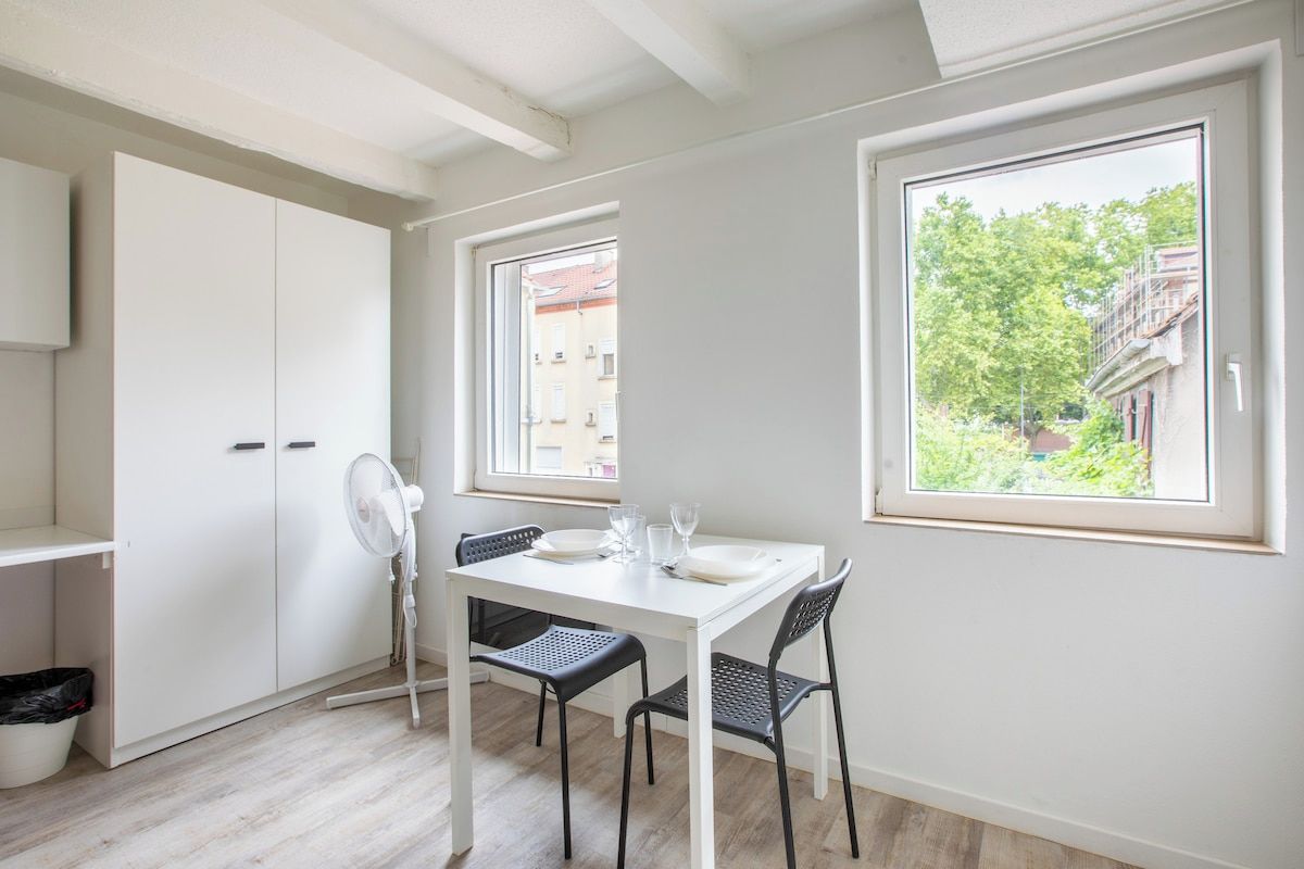 Una habitación con una mesa blanca, dos sillas, un ventilador y una ventana que ofrece vistas a edificios y árboles.