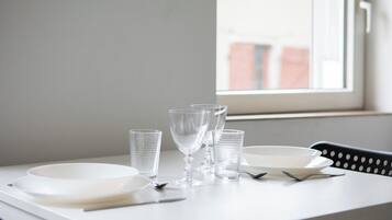 Una mesa dispuesta con platos blancos, vasos transparentes y cubiertos, ubicada cerca de una ventana con vistas a edificios.
