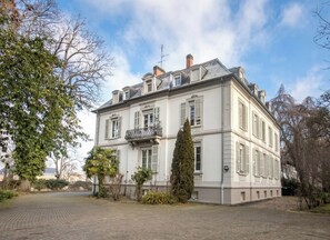 Een groot, twee verdiepingen hoog gebouw met een symmetrische gevel, meerdere ramen met luiken en een centraal balkon.