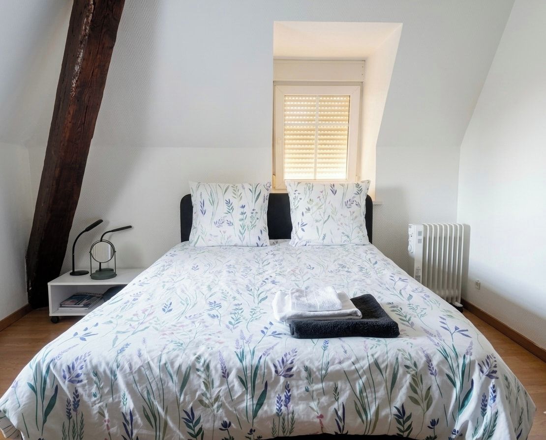 Ein Schlafzimmer mit einem Bett, einem Fenster mit Jalousien, einem Heizkörper und einem kleinen Regal mit Lampe und Büchern.