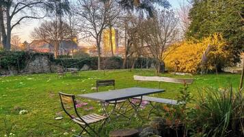 A grassy outdoor area with a table and chairs, surrounded by trees and a stone wall.