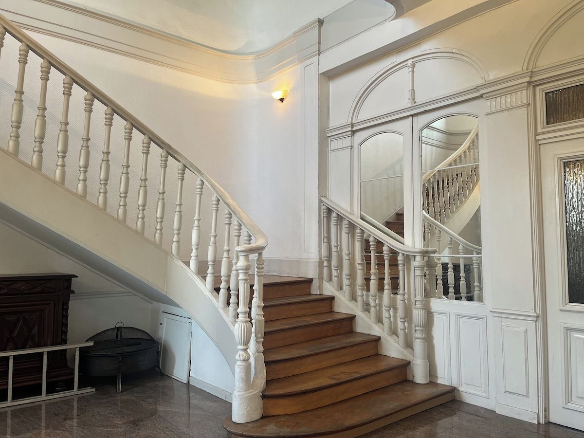 Un magnifique escalier avec des balustrades blanches et des marches en bois, menant à un palier surmonté d’une grande porte en arc de cercle.