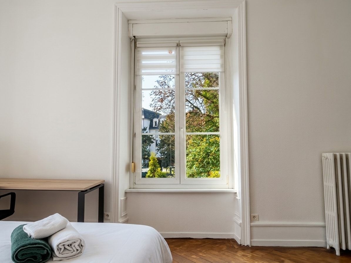 Ein Zimmer mit einem Bett, einem Fenster mit Jalousien, einem Heizkörper und Blick auf Bäume und ein Haus.