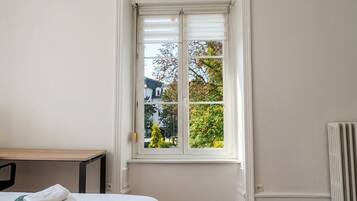 Ein Zimmer mit einem Bett, einem Fenster mit Jalousien, einem Heizkörper und Blick auf Bäume und ein Haus.