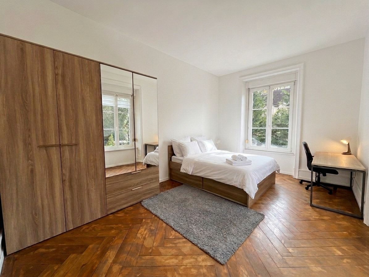 Une chambre à coucher comprenant un lit, un bureau, un miroir et un parquet en bois à motifs en chevrons.