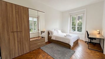 A bedroom with a bed, a desk, a mirror, and a herringbone-patterned wooden floor.