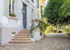 Een straat met keien leidt naar een gebouw met een trap en een bloembak met roze bloemen.