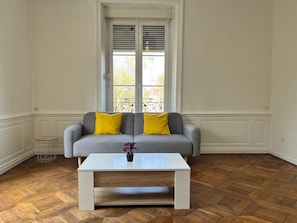 Een kamer met een grijze bank met gele kussens, een witte salontafel en een visgraat parketvloer.