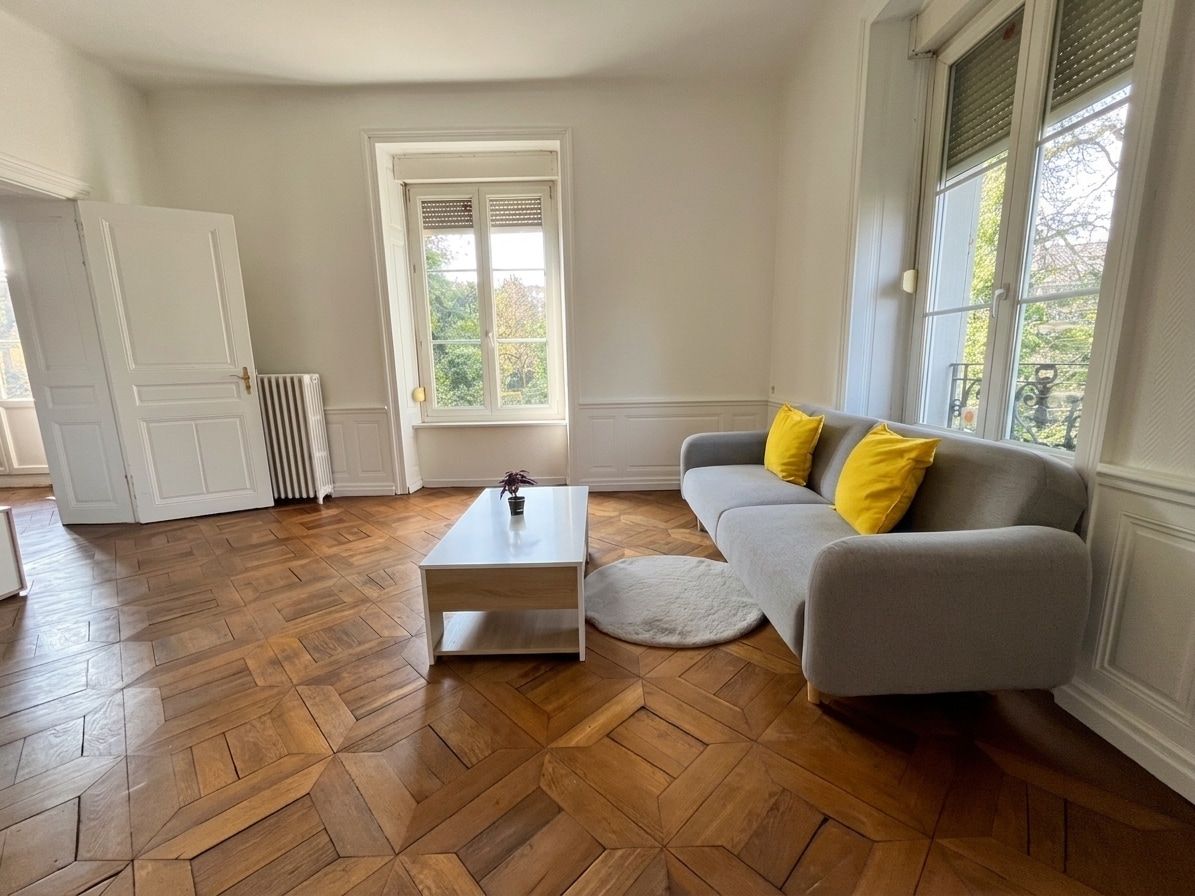 Un salon doté d’un parquet en bois à motifs chevrons, d’un canapé gris agrémenté de coussins jaunes, d’une table basse blanche et d’un radiateur.