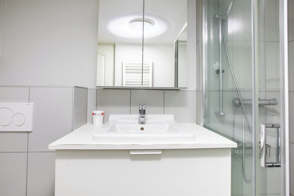 Un baño moderno con un lavamanos blanco, un espejo encima y una zona de ducha con mampara de vidrio.