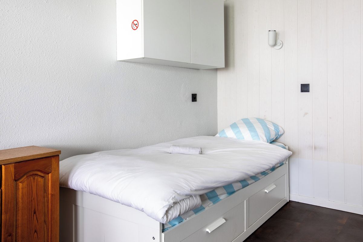 Un lit simple avec un matelas blanc et un oreiller à rayures bleues, une table de chevet en bois, une armoire blanche et un luminaire fixé au mur.
