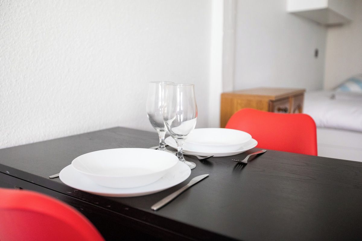 Una mesa de comedor dispuesta con platos blancos, cubiertos y dos copas de vino vacías.
