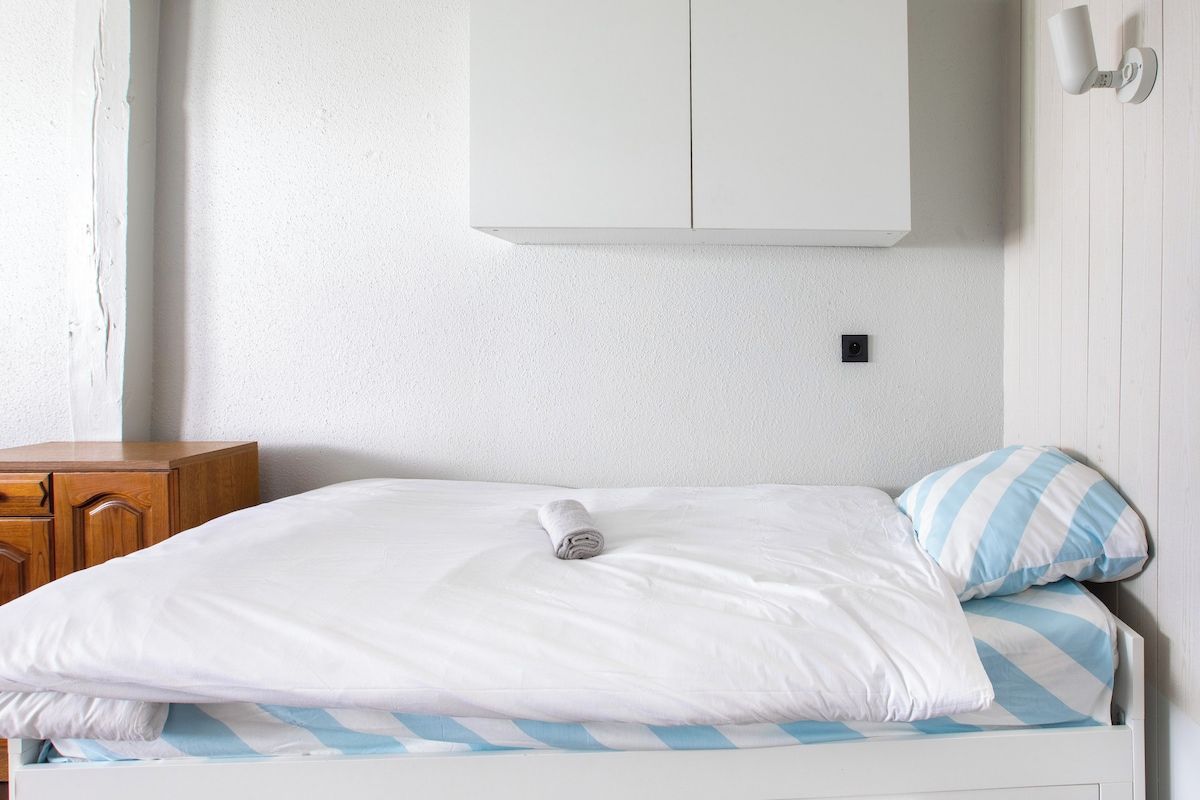 Un lit simple avec une literie blanche et un oreiller à rayures, une commode en bois et une armoire blanche placée au-dessus du lit.
