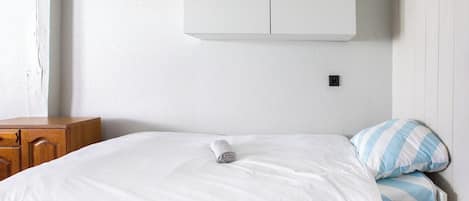 Een enkel bed met witte beddengoed en een gestreept kussen, een houten dressoir en een witte kast boven het bed.
