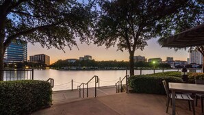 A riverside patio with a view of the city skyline, a wooden table and chairs, and a chain-link fence.
