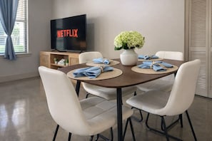 A dining area with a table set for four, a vase of flowers, and a television displaying the Netflix logo. - Lake Carolyn Std Near Toyota MusicFactory Pool+GYM (Irving)