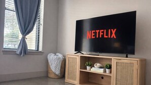 Una stanza con televisione che mostra 'NETFLIX', una finestra con tende, un cuscino a forma di cuore e un mobiletto in legno con oggetti decorativi.