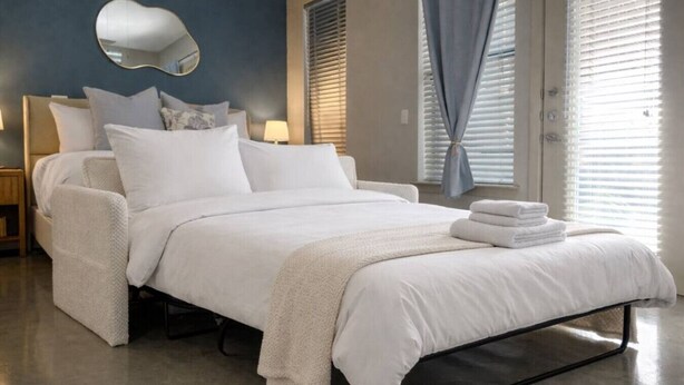 A modern bedroom with a bed, pillows, a bedside table, a lamp, and a window with blinds.